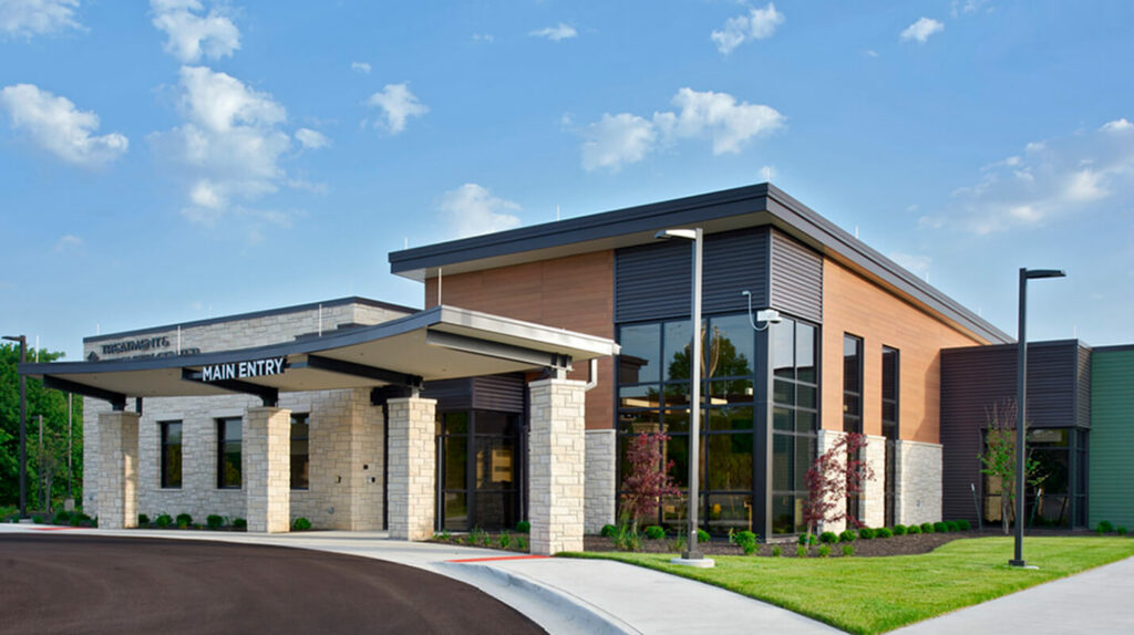 A wide-angle view of the Douglas County Crisis Center, capturing the full building and its surroundings. The building has a sleek, modern design with large glass panels and a neutral color palette. The landscape is neatly maintained, with green lawns and a few trees providing a serene environment.