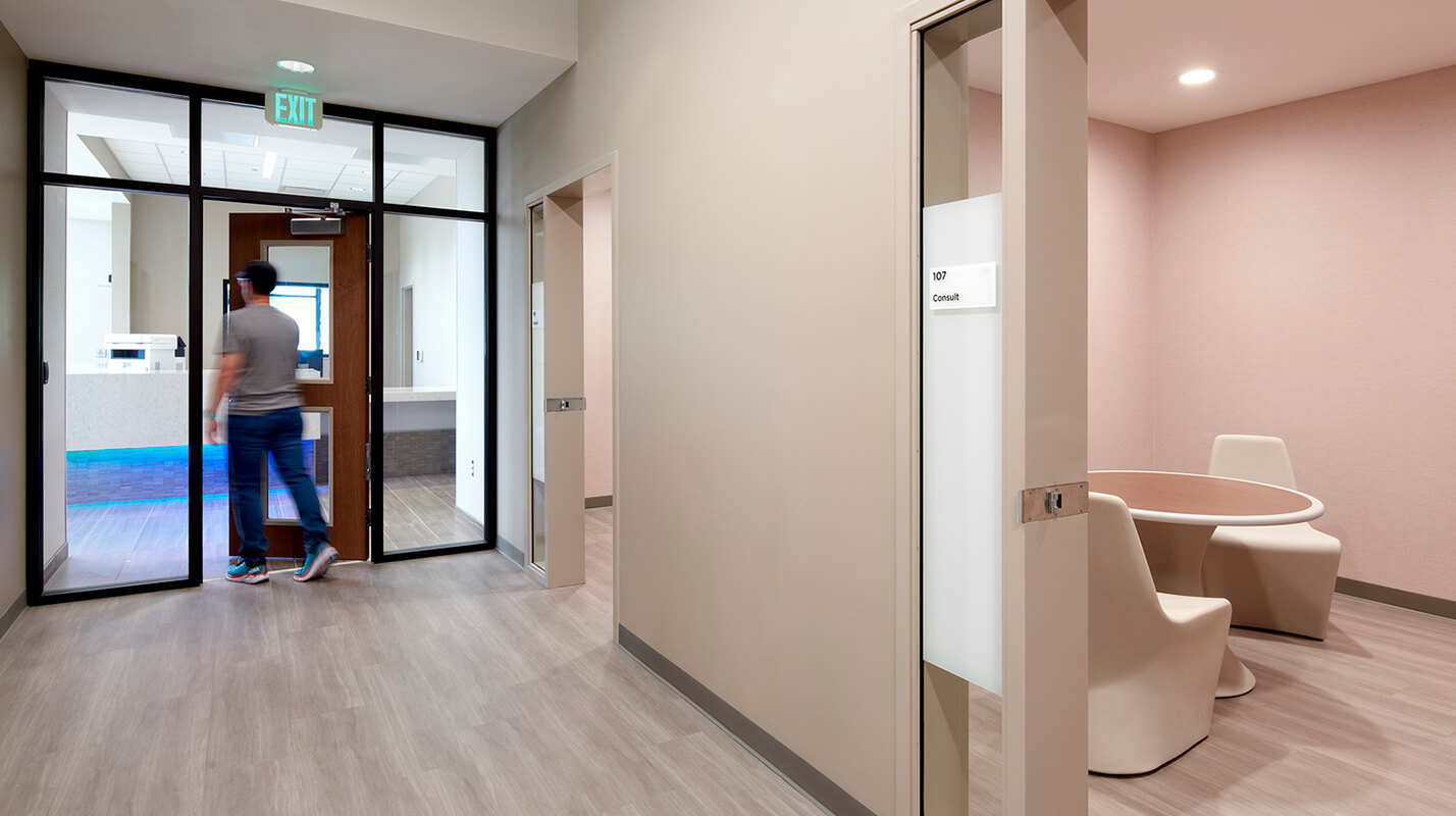 An interior hallway of the Douglas County Crisis Center, featuring a long corridor with light-colored walls and a polished floor. The design is clean and minimalist, with large windows along one side of the hallway allowing natural light to fill the space. The hallway leads to various rooms, each marked with clear signage.