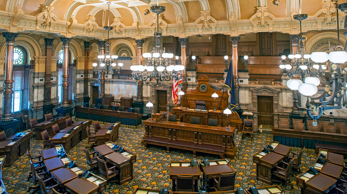 Photo shows the Kansas Senate chambers, c2013, after the room was resorted. It features the historically appropriate patterned carpet, decoratively painted walls, historic desks, historic gallery, and 28 restored copper columns.