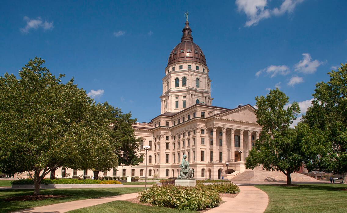 Photo of the Kansas Statehouse, a grand building with a dome, with a statue in the foreground.