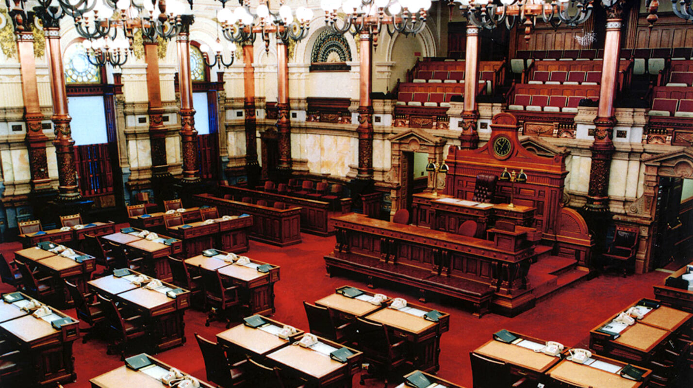 Photo of the Kansas Senate chambers, circa 1999, with historic senate desks and gallery seating. The photo is intended to demonstrate the non-historic elements such as the red carpet, cream wall paint, and chandeliers.