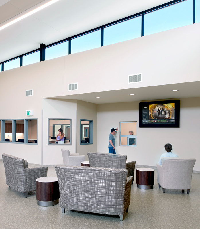 A modern, minimalist interior of a crisis center, featuring a spacious room with large windows allowing ample natural light. The room is furnished with a few simple chairs arranged around a coffee table, with light-colored wooden floors and white walls. The atmosphere is calm and welcoming.