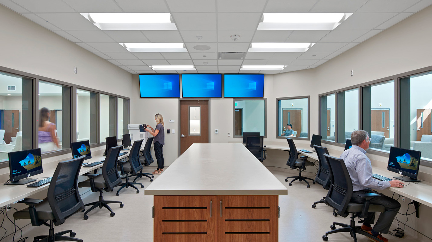 A modern observation room with workstations, large windows for visibility, and bright lighting. People are seen working and moving around.