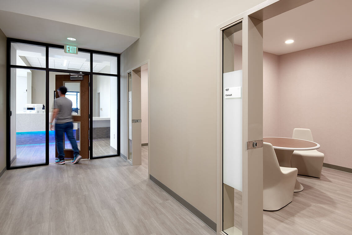 An interior hallway of the Douglas County Crisis Center, featuring a long corridor with light-colored walls and a polished floor. The design is clean and minimalist, with large windows along one side of the hallway allowing natural light to fill the space. The hallway leads to various rooms, each marked with clear signage.