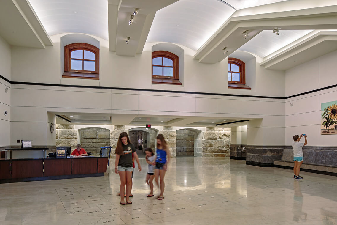 Photo shows children standing on a map of Kansas and its counties with a woman sitting at a desk in the background in the Kansas Statehouse visitor center.