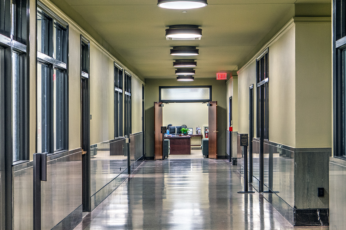 A long, polished corridor with glass office partitions and modernized overhead lighting. This careful rehabilitation retains original proportions and finishes while improving lighting and visibility for everyday academic use.
