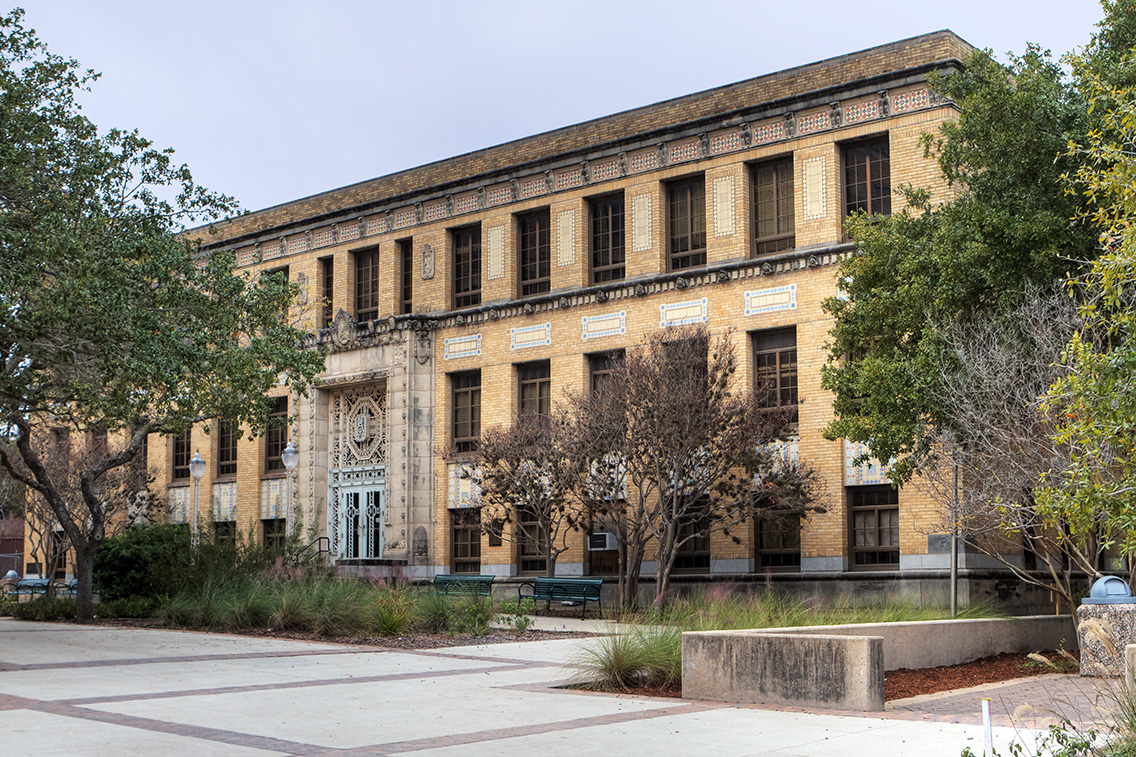 An oblique view of Scoates Hall, a three-story yellow brick academic building featuring detailed terra cotta ornamentation, symmetrical windows, and a central, formal entry framed by decorative ironwork. The well-maintained landscape and mature trees reinforce its role as a historic campus cornerstone.