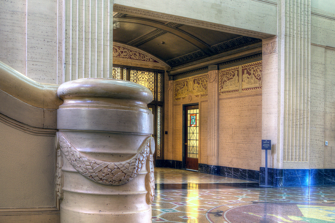 Close-up of a polished balustrade post and corridor view revealing bas-relief murals, ornate metalwork, and vaulted ceilings. The craftsmanship highlights the care taken in both public and transitional spaces, honoring the building’s civic legacy.