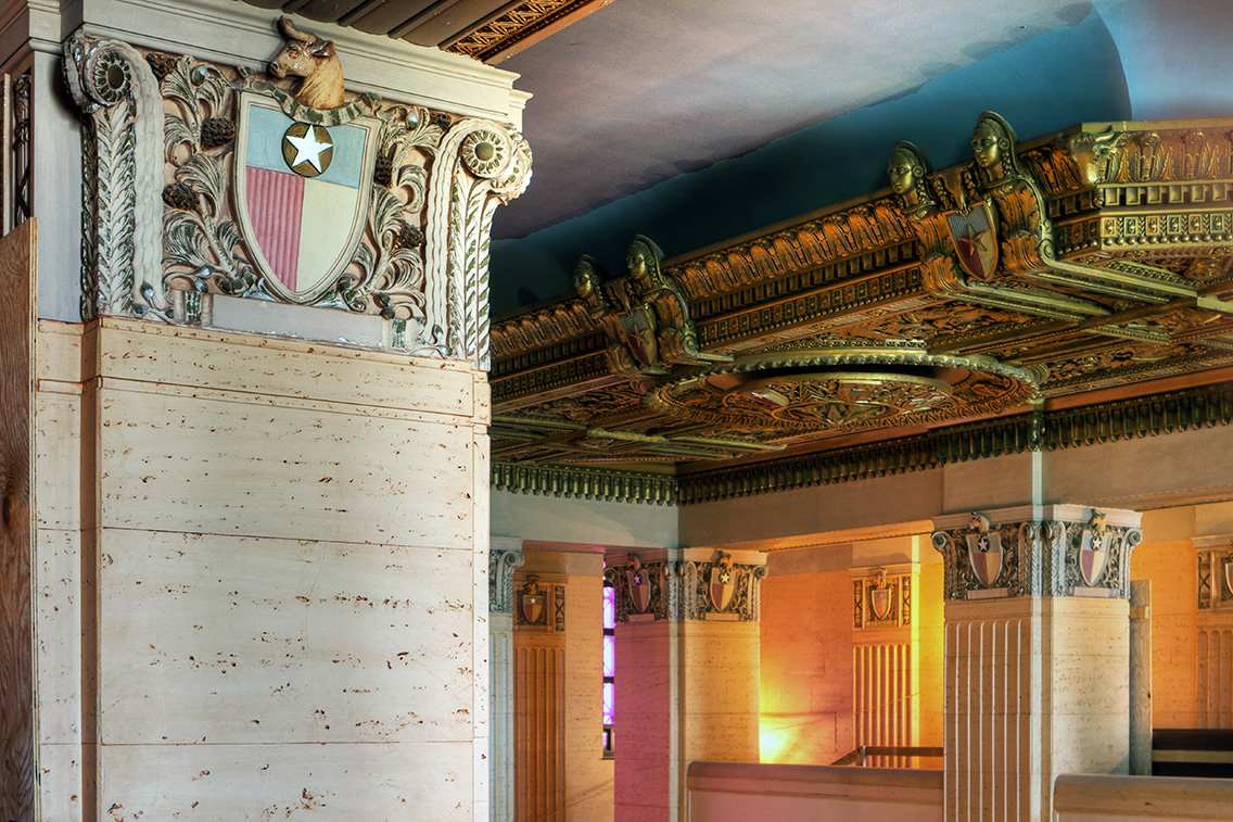 Close-up of a decorative capital and a vibrant gilded coffered ceiling. Painted shields, sculptural figures, and a bull’s head motif exemplify narrative ornamentation, each detail speaking to the building’s cultural role and identity.