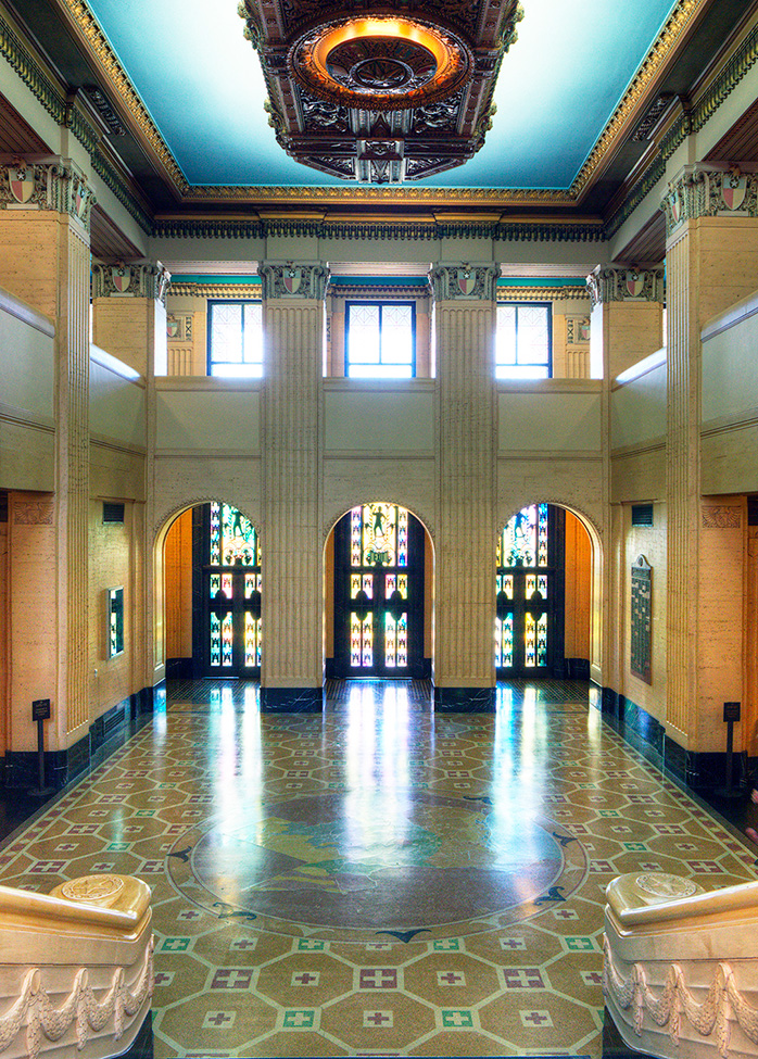 A double-height public hall framed by monumental columns and light-washed travertine. The stained-glass doors and floor medallion map root the space in both time and place, offering visitors a powerful sense of civic grandeur and orientation.