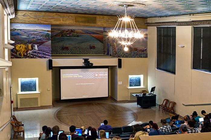 Students in a historic Texas A&M lecture hall with murals and decorative ceiling.
