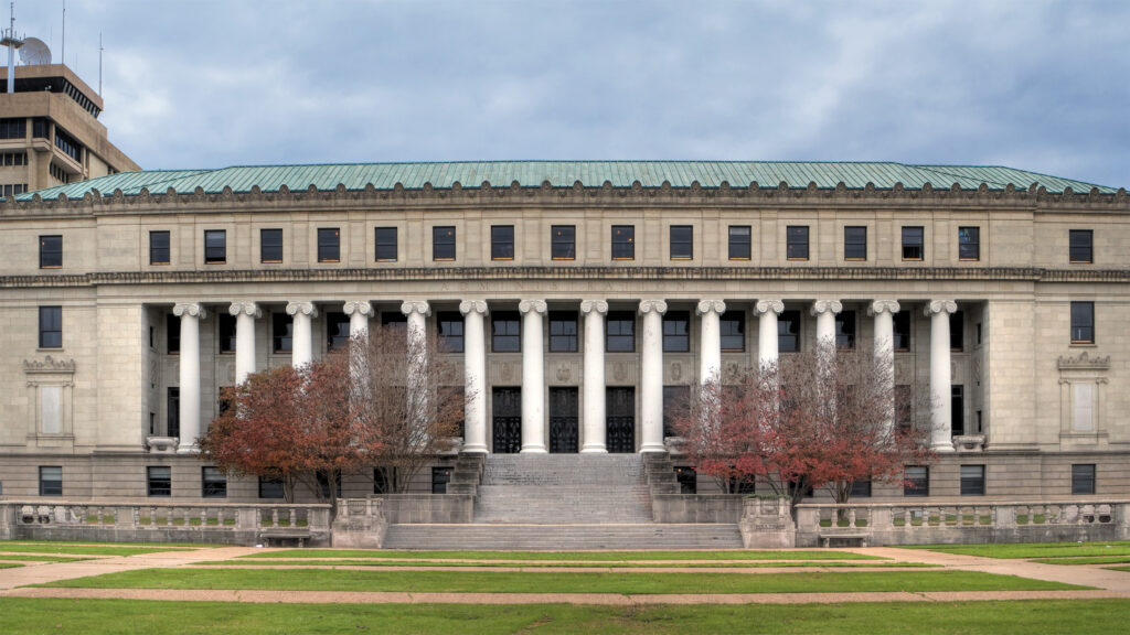 Straight-on view of a monumental neoclassical building featuring a rhythm of Ionic columns, a limestone façade, and a copper roof. The symmetry, balanced by seasonal trees lining the entry stair, captures the enduring civic presence and formal dignity of the structure.